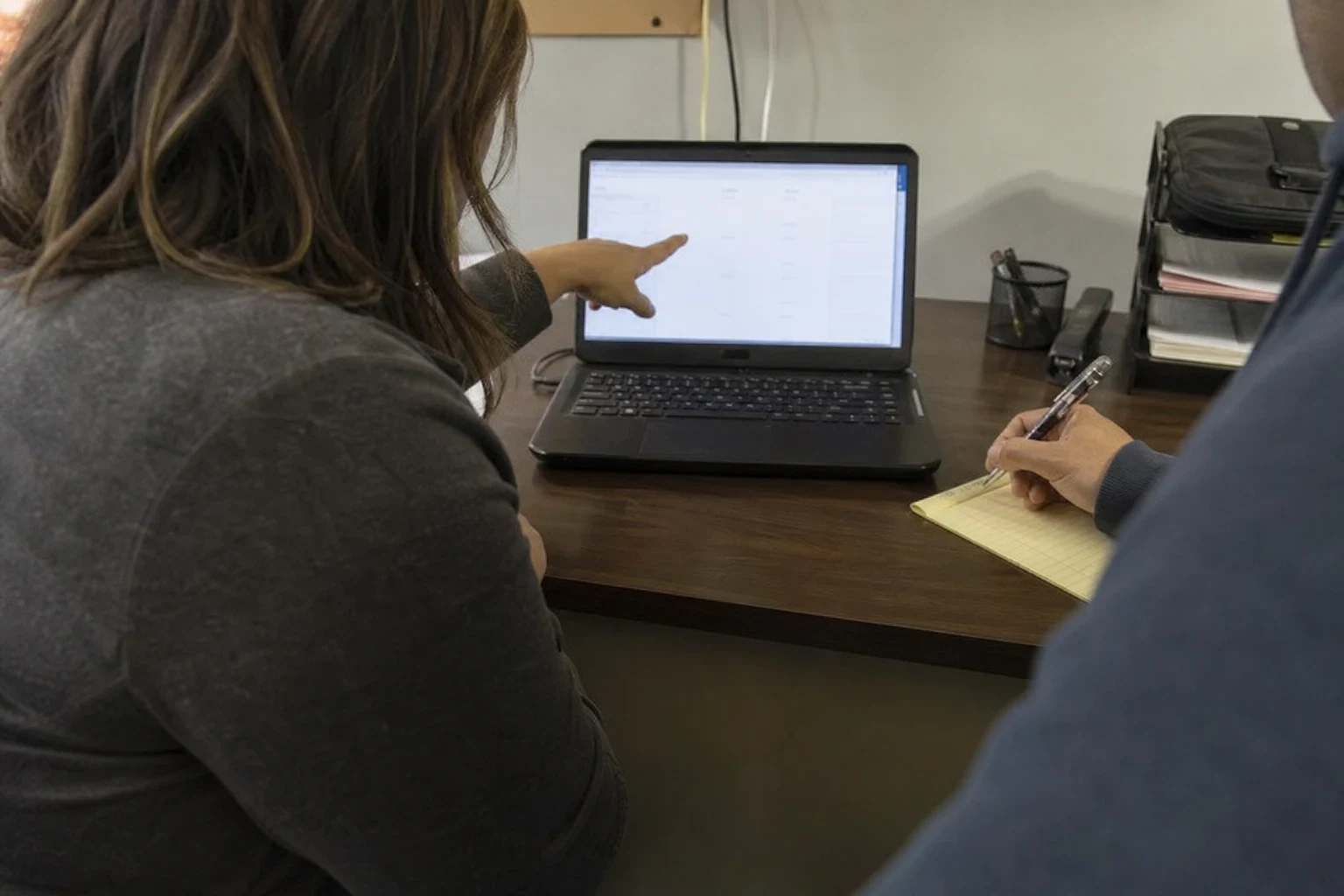 Over-the-shoulder view of a woman pointing at a laptop screen on an office desk while a man beside her takes notes on a yellow legal pad