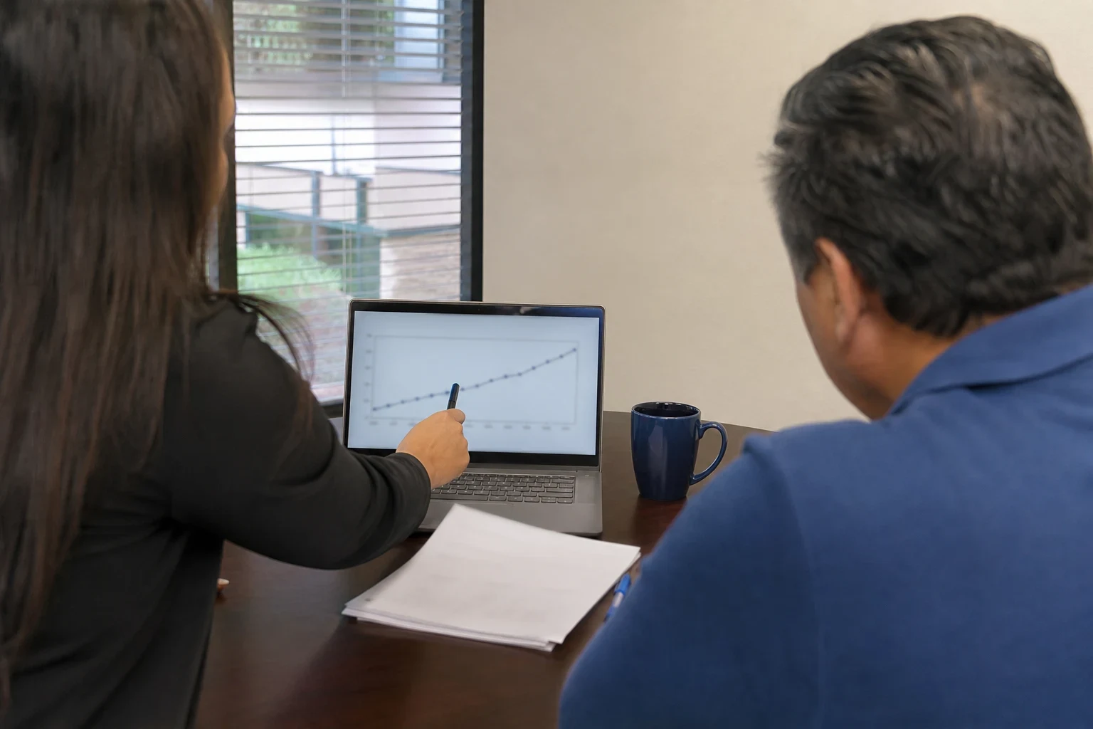 Agent points a pen at a rising line chart on a laptop while a client in a blue shirt looks on at an office desk with papers, a navy mug and a window with blinds
