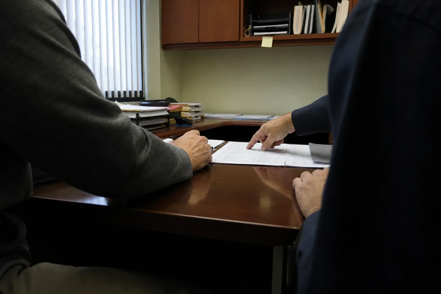 Two people seated at a wooden office desk, one pointing at printed documents while the other holds a pen, with shelves and window blinds behind