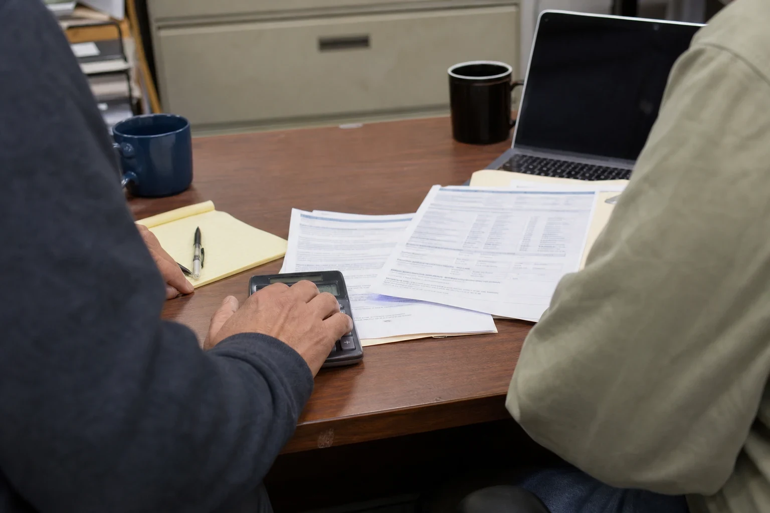 Two people at a wooden office desk reviewing printed statements, one tapping a calculator beside a yellow legal pad, with mugs, a laptop and a filing cabinet behind